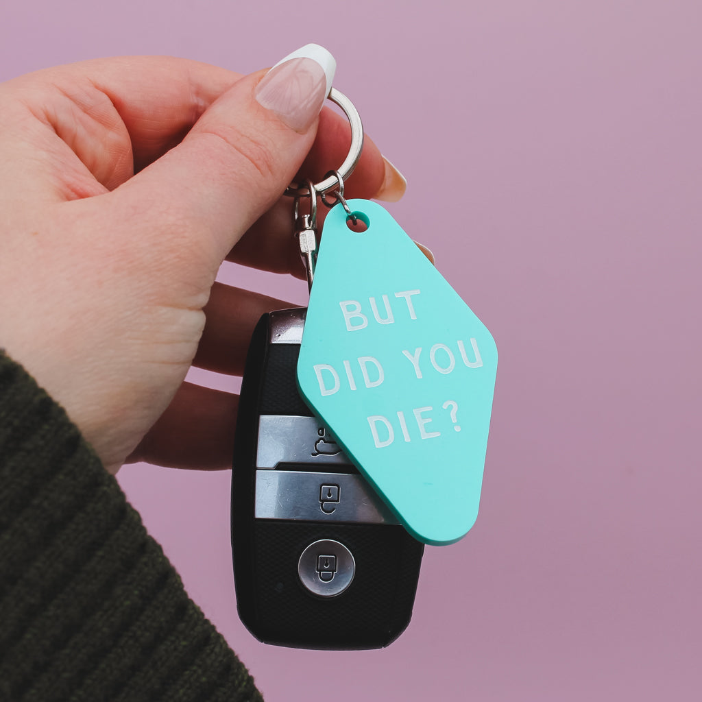 Motel style keychain with 'But did you die?' tag held by a hand against a pink background
