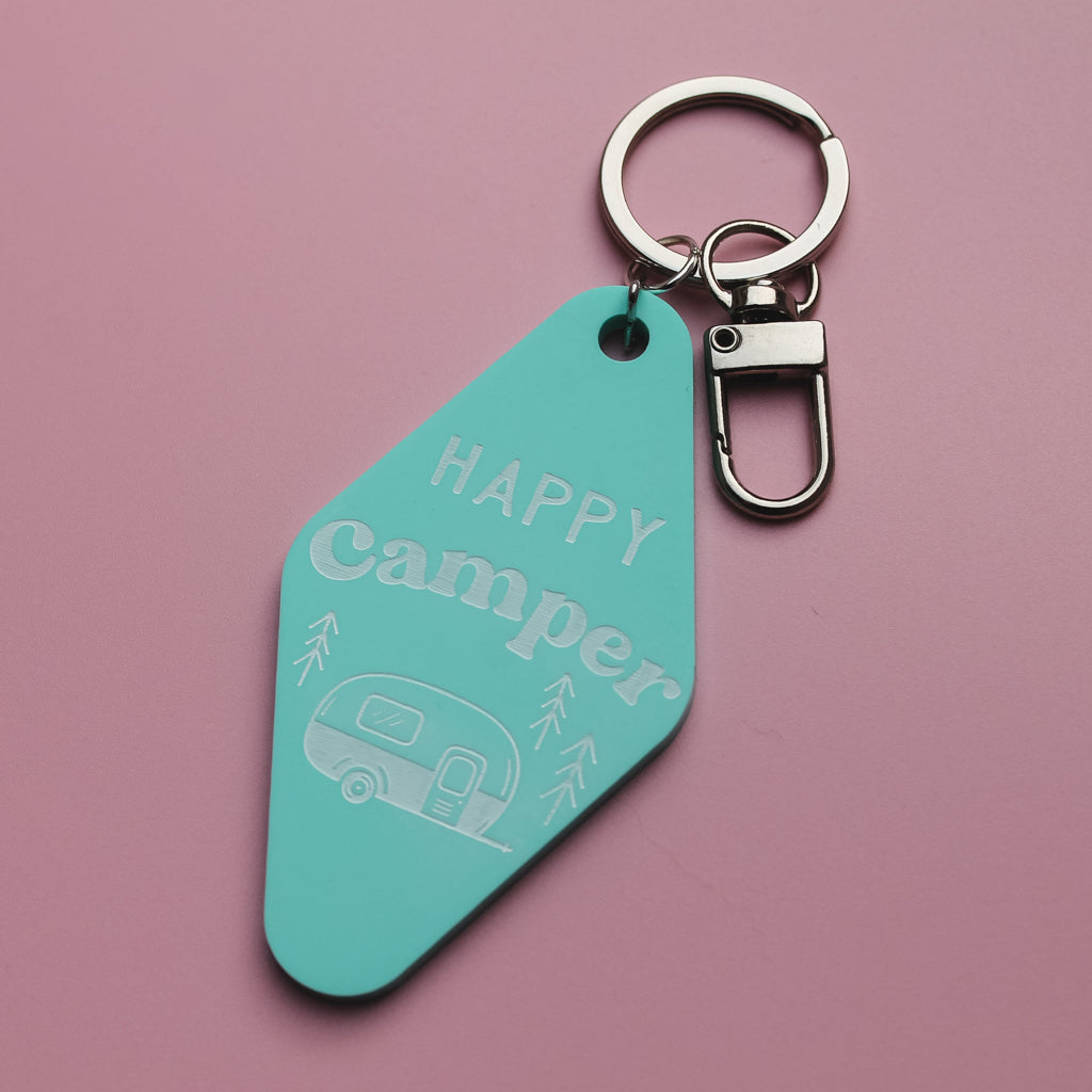 Turquoise motel style keychain with 'Happy Camper' text and caravan illustration on a pink background