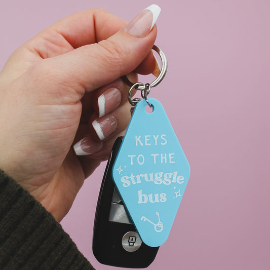 Hand holding a keychain with a blue tag reading 'Keys to the Struggle Bus' against a pink background