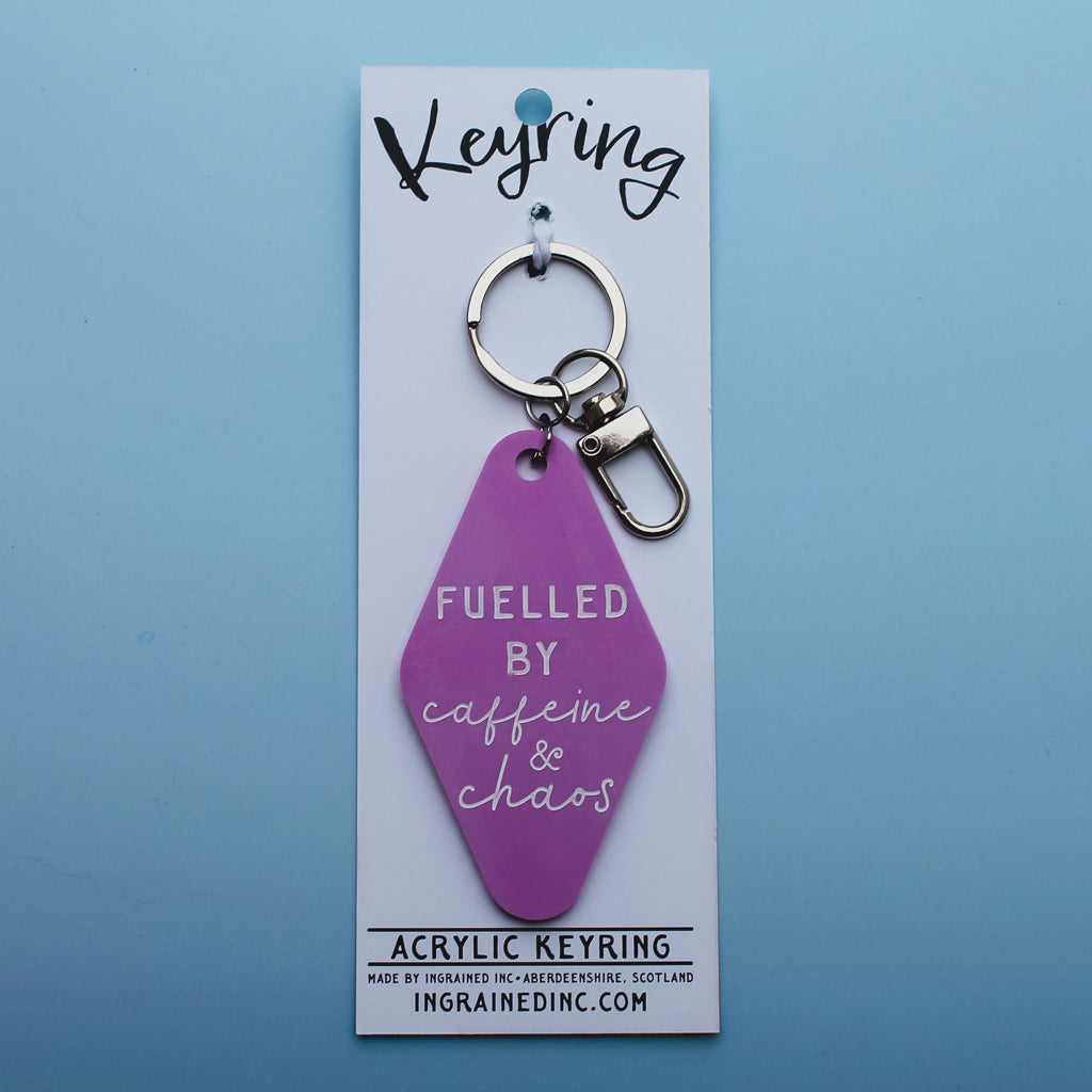 Pink Motel style keyring with 'Fuelled by caffeine & chaos' text in its packaging on a blue background