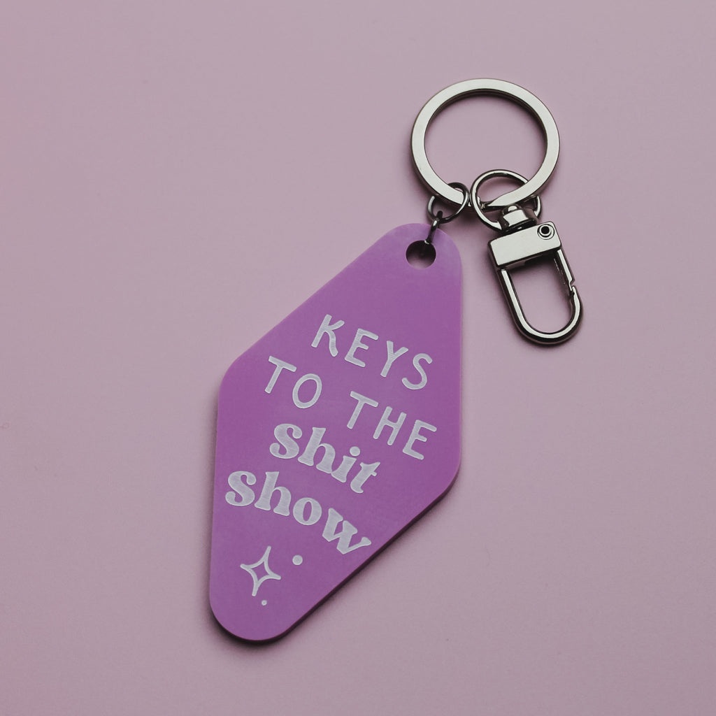 Purple motel style keychain saying keys to the shit show with white text on a pink background
