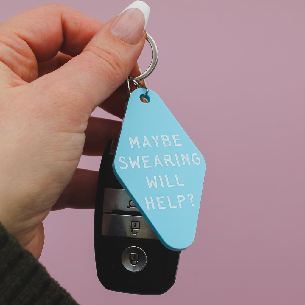 Hand holding a blue motel style keychain saying 'MAYBE SWEARING WILL HELP?' against a pink background