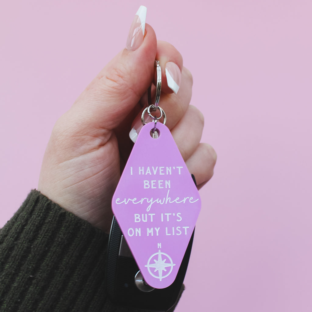 Hand holding a pink motel style keychain with the quote 'I haven't been everywhere, but it's on my list' against a pink background