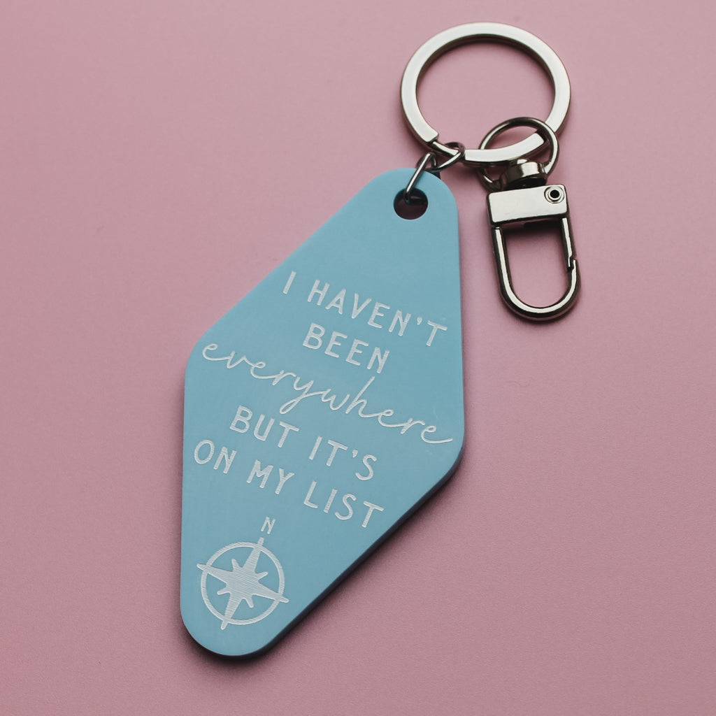 Blue motel style keychain with the quote 'I haven't been everywhere, but it's on my list'  compass design on a pink background