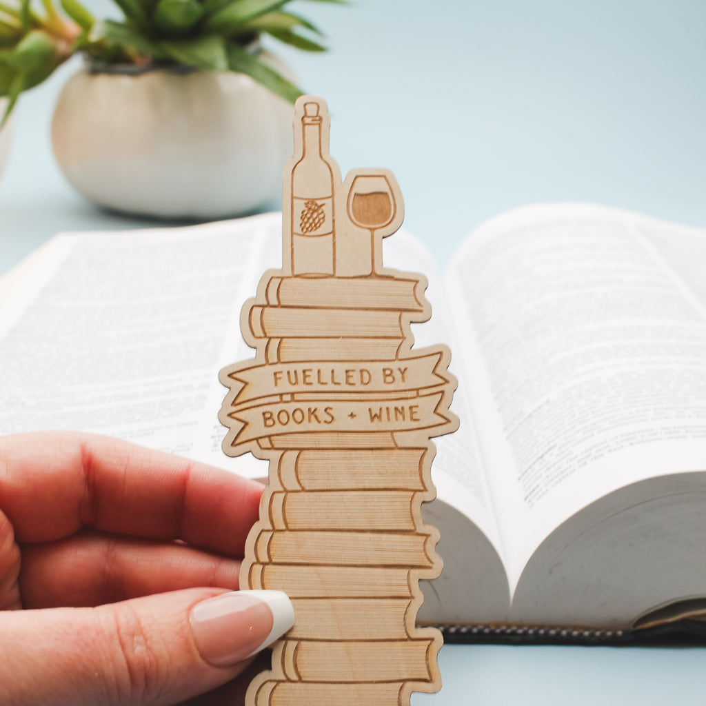 Wooden bookmark shaped like a wine bottle with 'Fuelled by Books + Wine' text, held over an open book.
