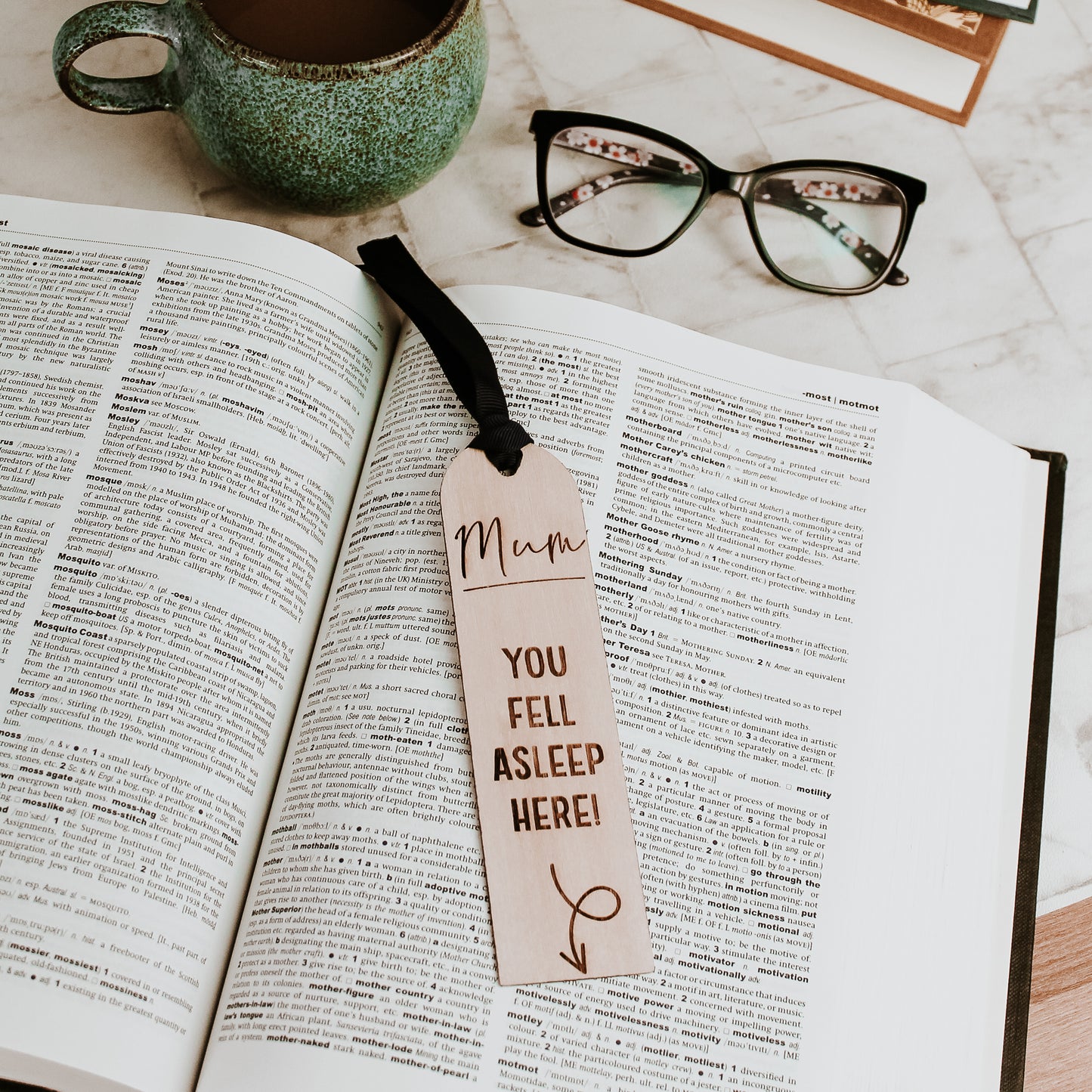 Personalized wooden bookmark engraved with "Mum, you fell asleep here" and an arrow pointing to the bottom. The bookmark rests on an open book with a navy blue ribbon attached.