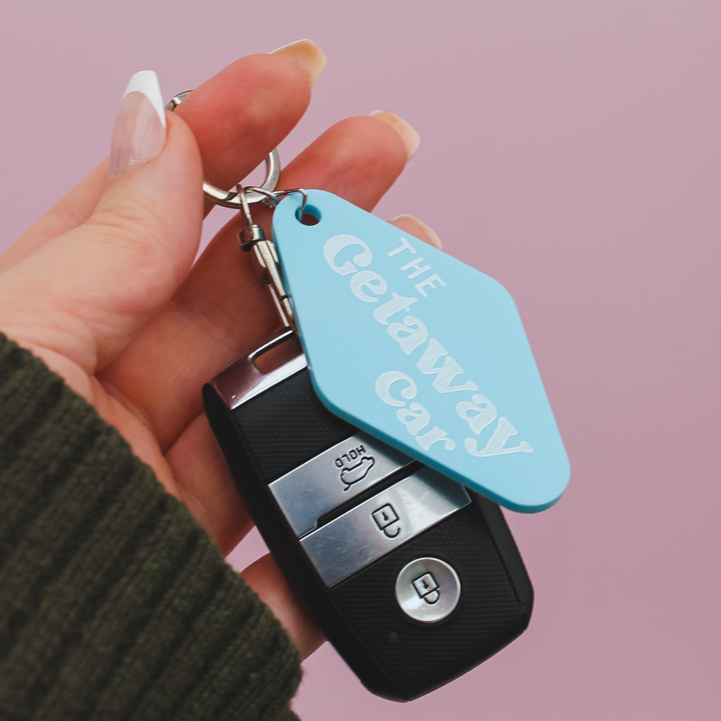 Hand holding a car key fob with a blue motel style keyring labelled 'The Getaway Car' against a pink background