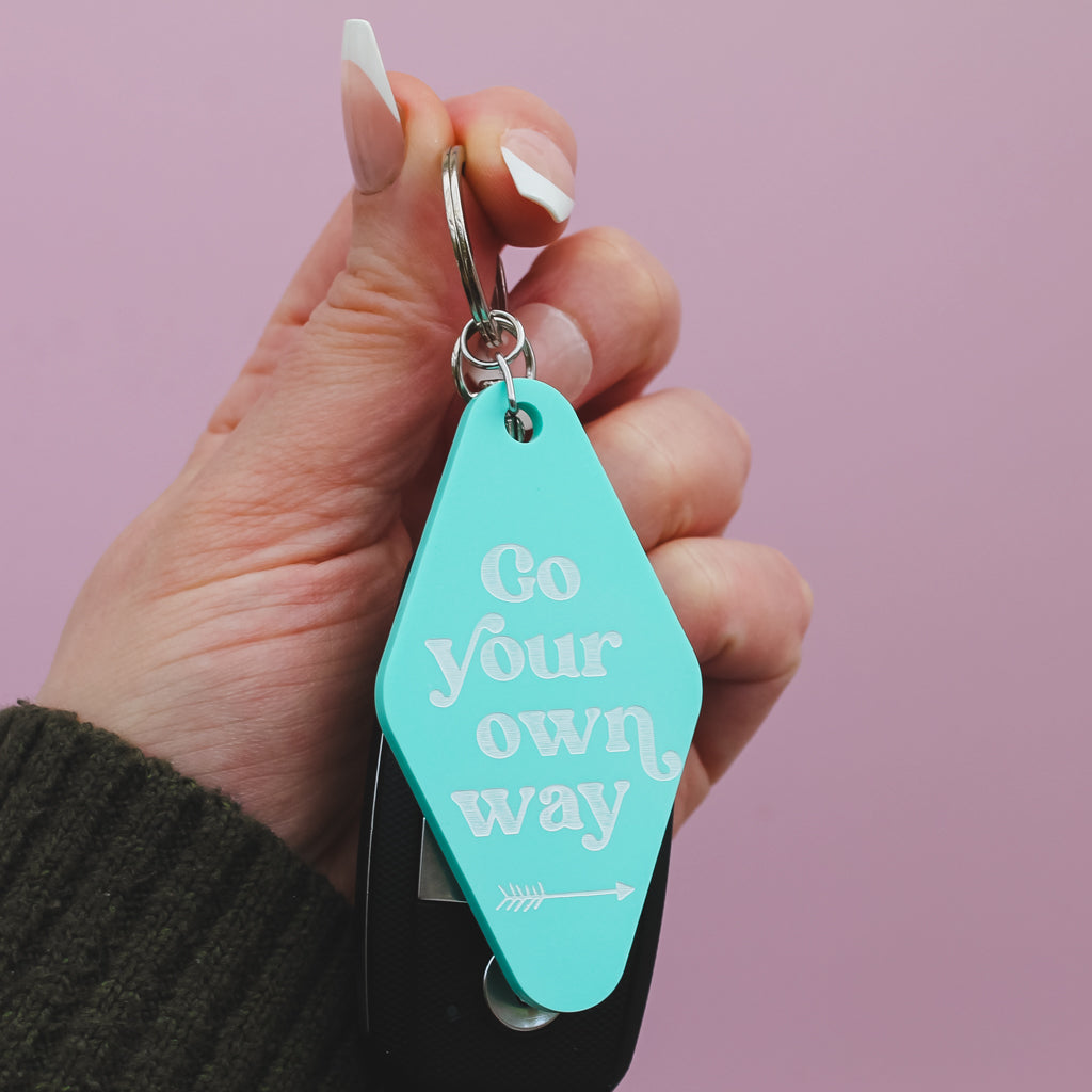 Hand holding a turquoise motel style keychain with 'Go your own way' text against a pink background