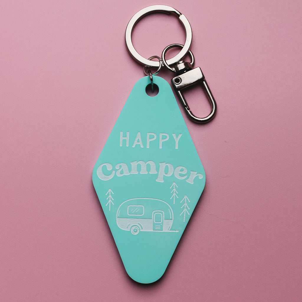 Turquoise motel style keyring with 'Happy Camper' text and caravan illustration on a pink background