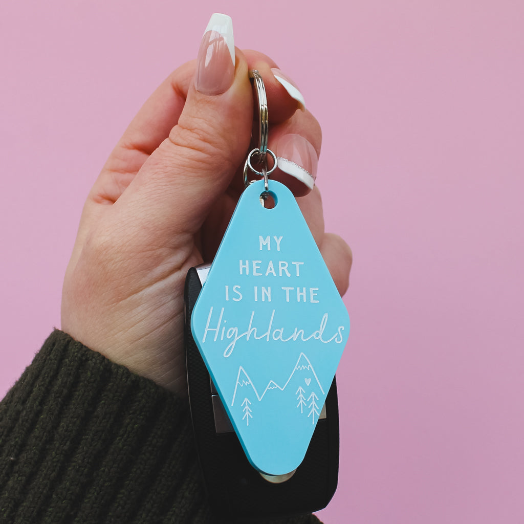 Hand holding a motel style keyring with a blue tag that says 'My heart is in the Highlands' against a pink background