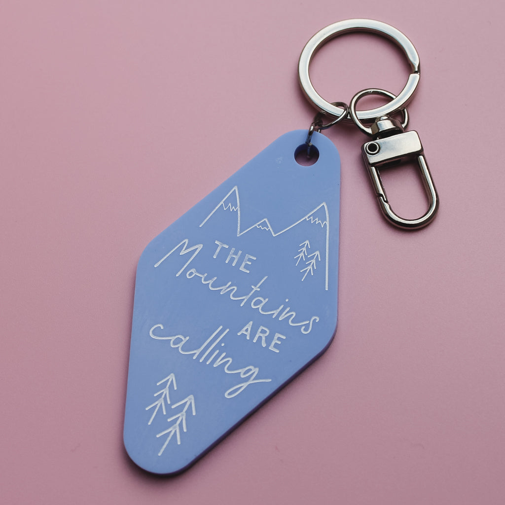 Blue motel style keyring with 'The Mountains Are Calling' text on a pink background