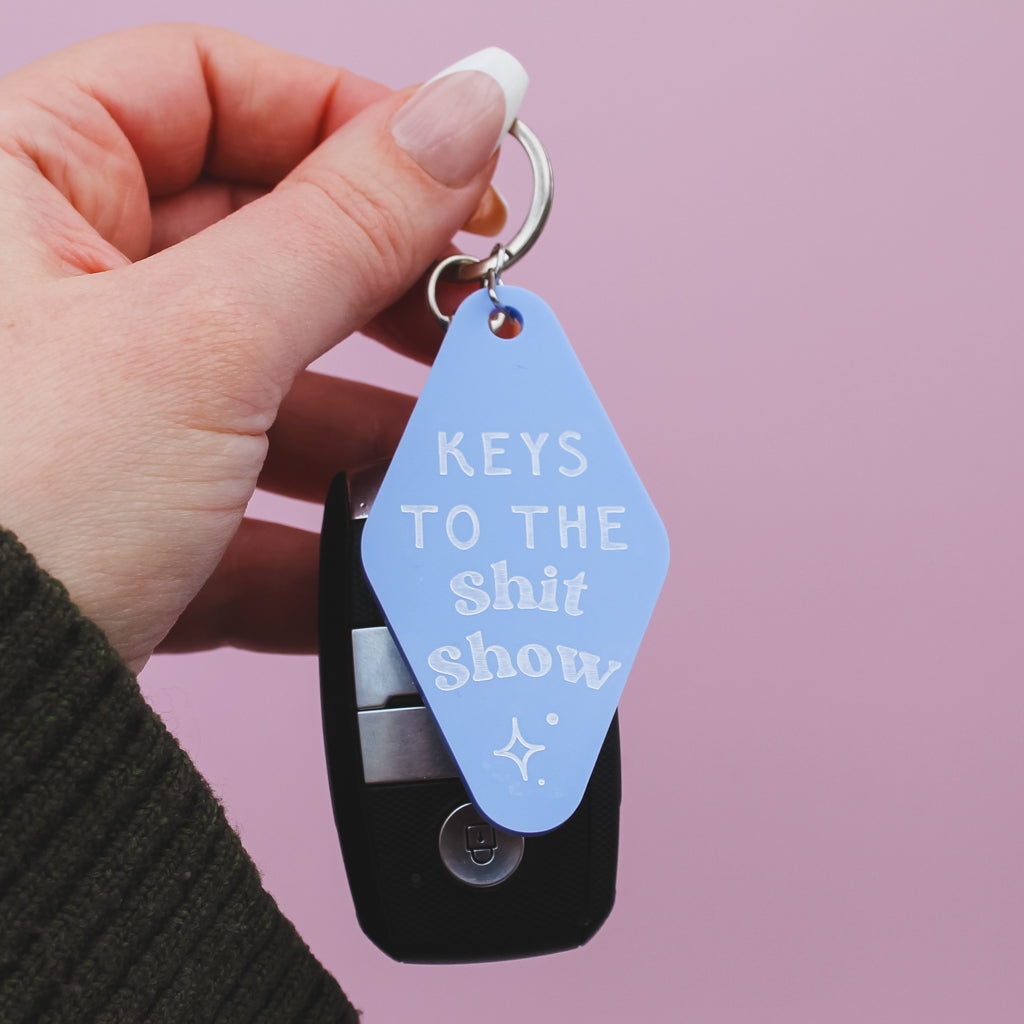 Hand holding a motel style keyring with 'Keys to the Shit Show' text against a pink background
