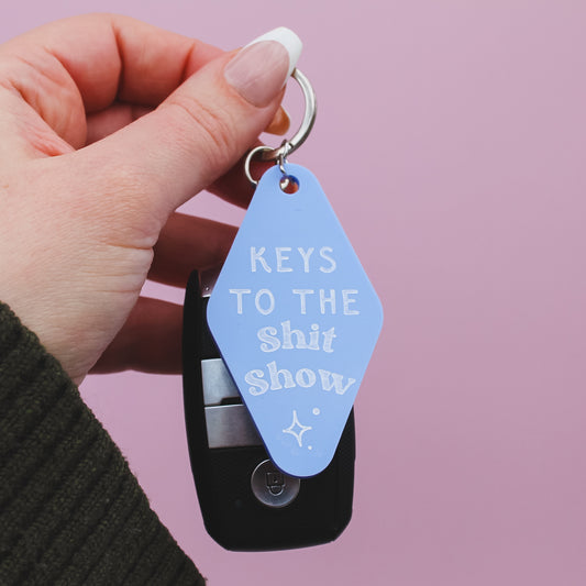 Hand holding a motel style keyring with 'Keys to the Shit Show' text against a pink background