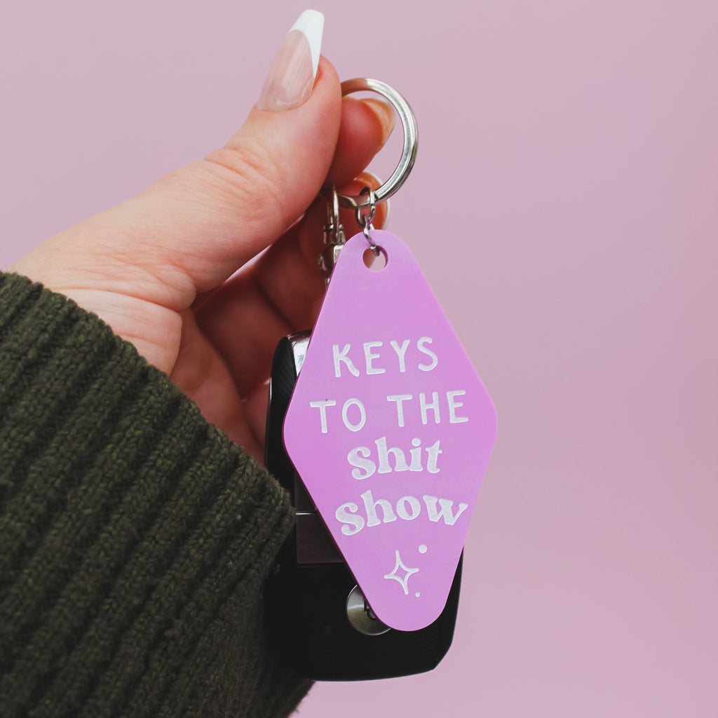 Hand holding a pink motel style keyring with white text reading 'keys to the shit show' against a pink background