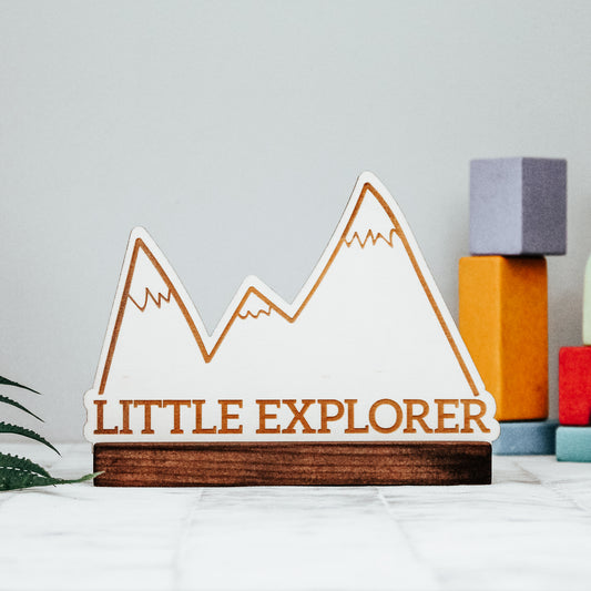 wooden standing mountain nursery decoration with the words little explorer engraved onto it in a scandi rustic aesthetic 