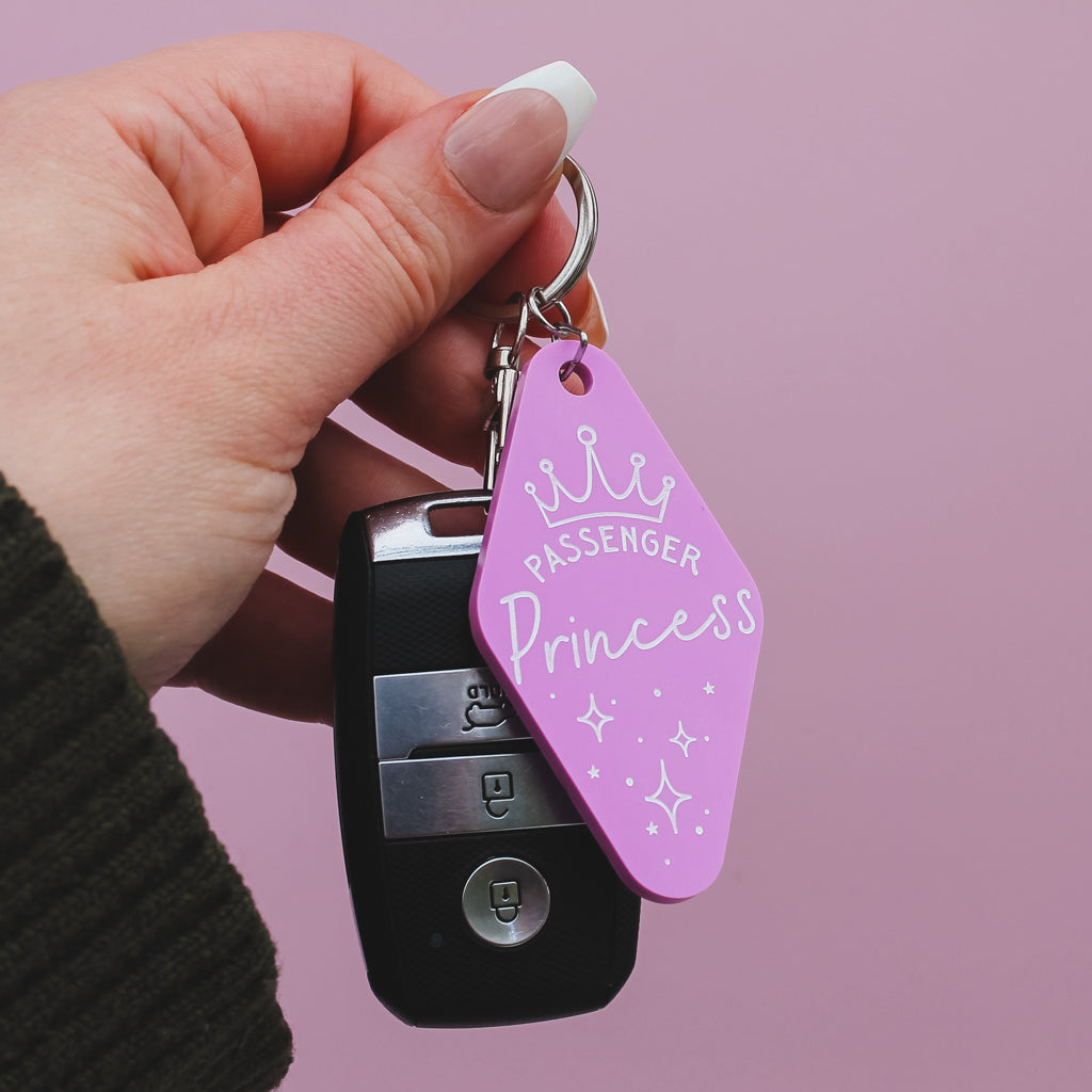 Hand holding a motel style keychain with a 'Passenger Princess' design on against a pink background