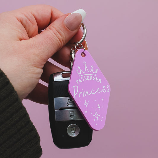 Hand holding a motel style keychain with a 'Passenger Princess' design on against a pink background