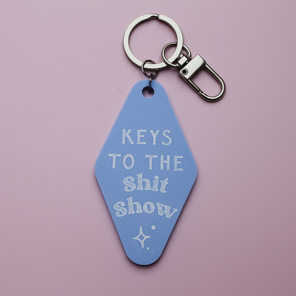 Blue motel style keychain tag with white text saying 'keys to the shit show' on a pink background
