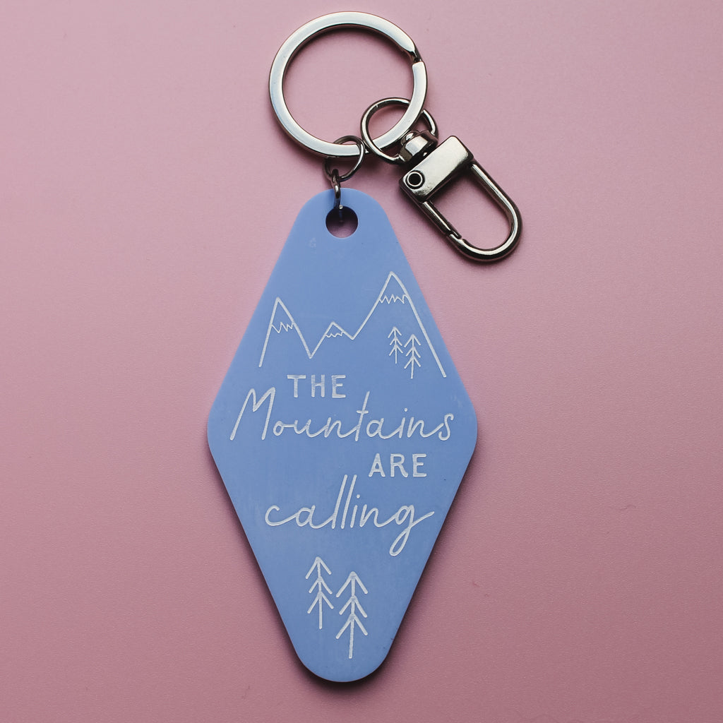 Blue motel style keychain with 'The Mountains Are Calling' text on a pink background