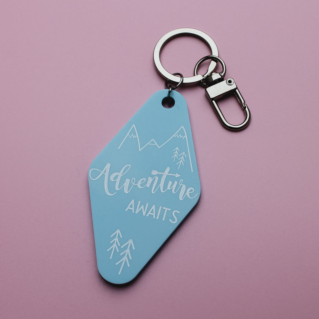 Blue motel style keyring tag with 'Adventure Awaits' text on a pink background