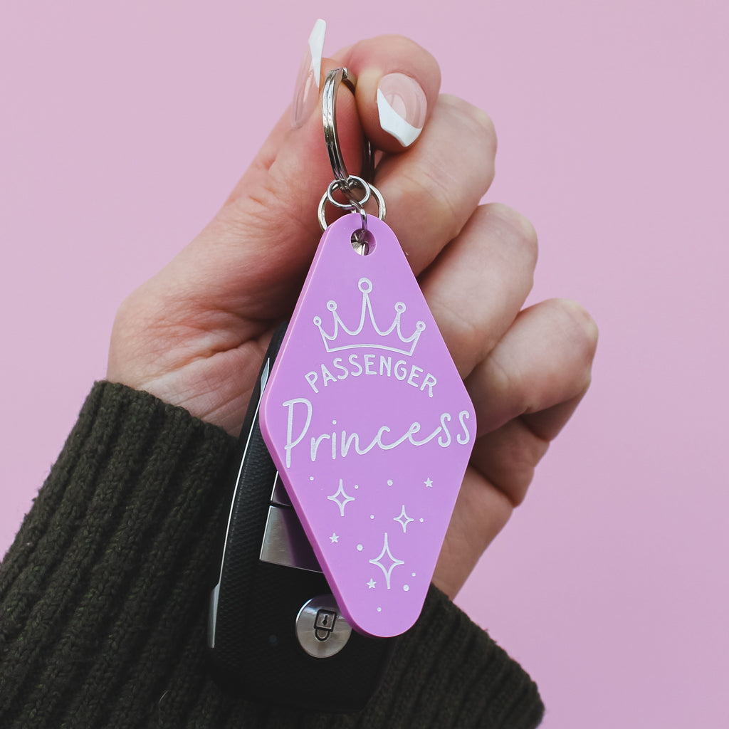 Hand holding a motel style pink keychain with 'Passenger Princess' tag against a pink background