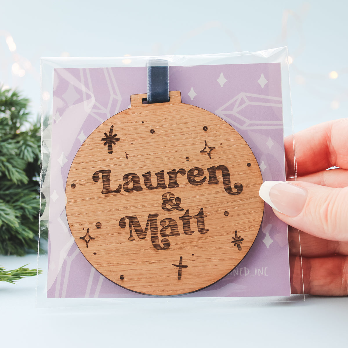 Wooden ornament with 'Lauren & Matt' engraving on a clear packaging with a light purple background.