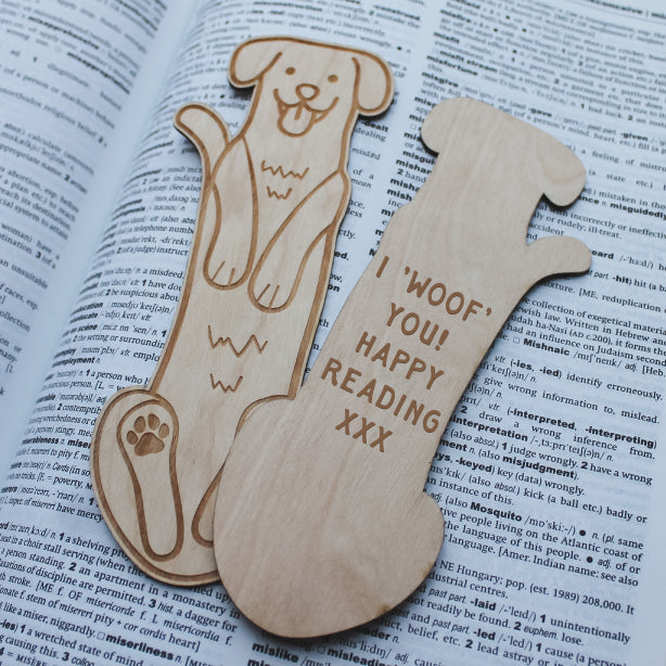 Wooden dog-shaped bookmarks with personalised text reading 'Woof You! Happy Reading XXX' on an open book.