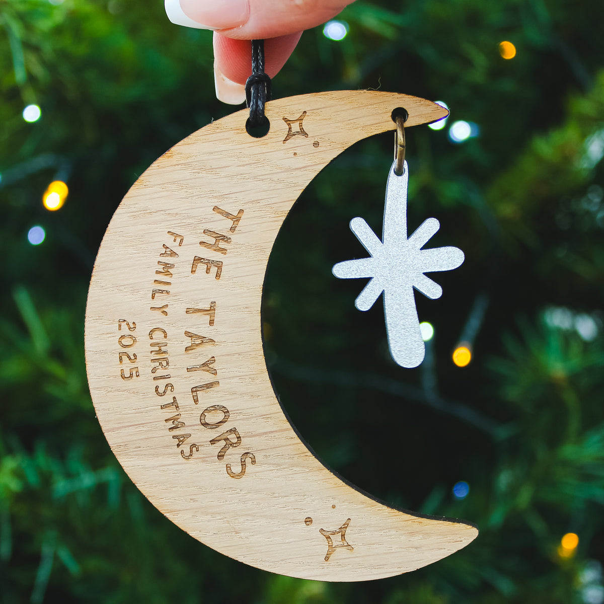 Wooden crescent moon ornament with personalised newlywed couple surname  engraved. Held against a Christmas tree background.