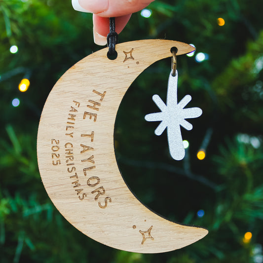 Wooden crescent moon ornament with personalised newlywed couple surname  engraved. Held against a Christmas tree background.