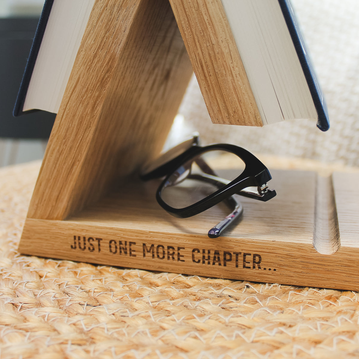A close-up of a solid oak wooden A-frame book stand engraved with the phrase 'JUST ONE MORE CHAPTER.