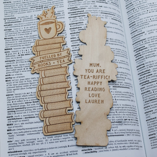 Two wooden bookmarks on an open book with one reading 'Fuelled by books & tea' and the other a personalized message.