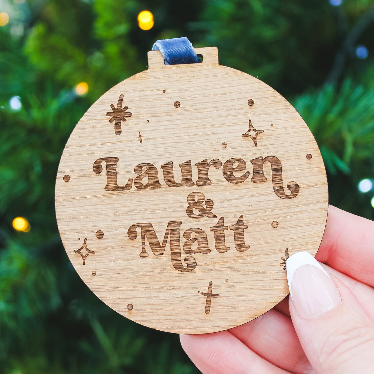 Wooden Christmas bauble ornament with personalised names engraved, held against a blurred green background.