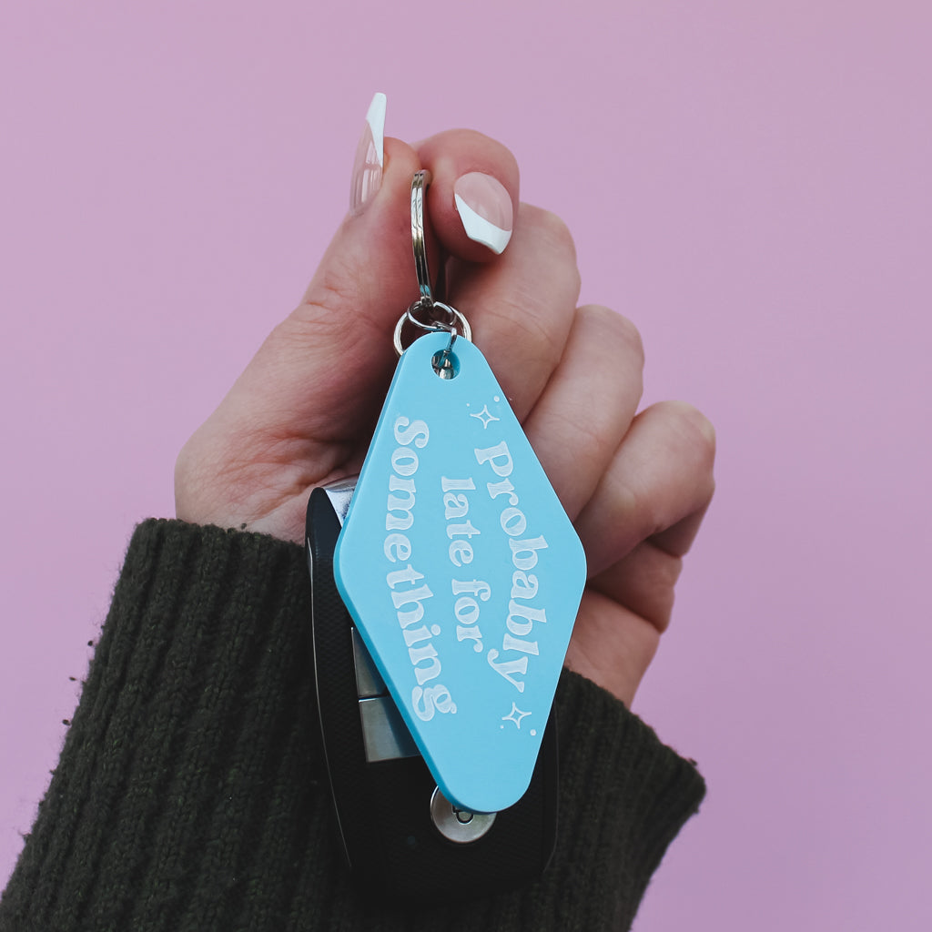 Hand holding a blue motel style keyring with text saying 'probably late for something' against a pink background