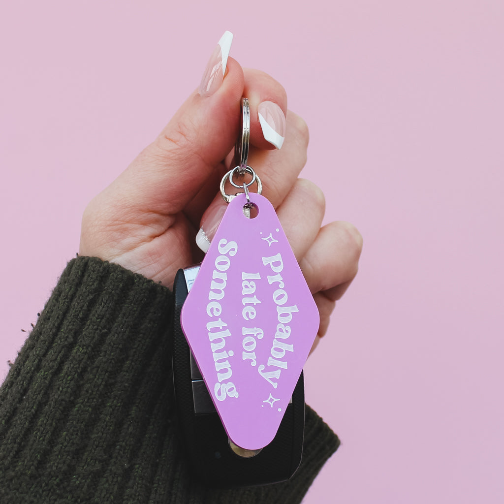 Hand holding a motel style pink keyring with text saying 'probably late for something' against a pink background