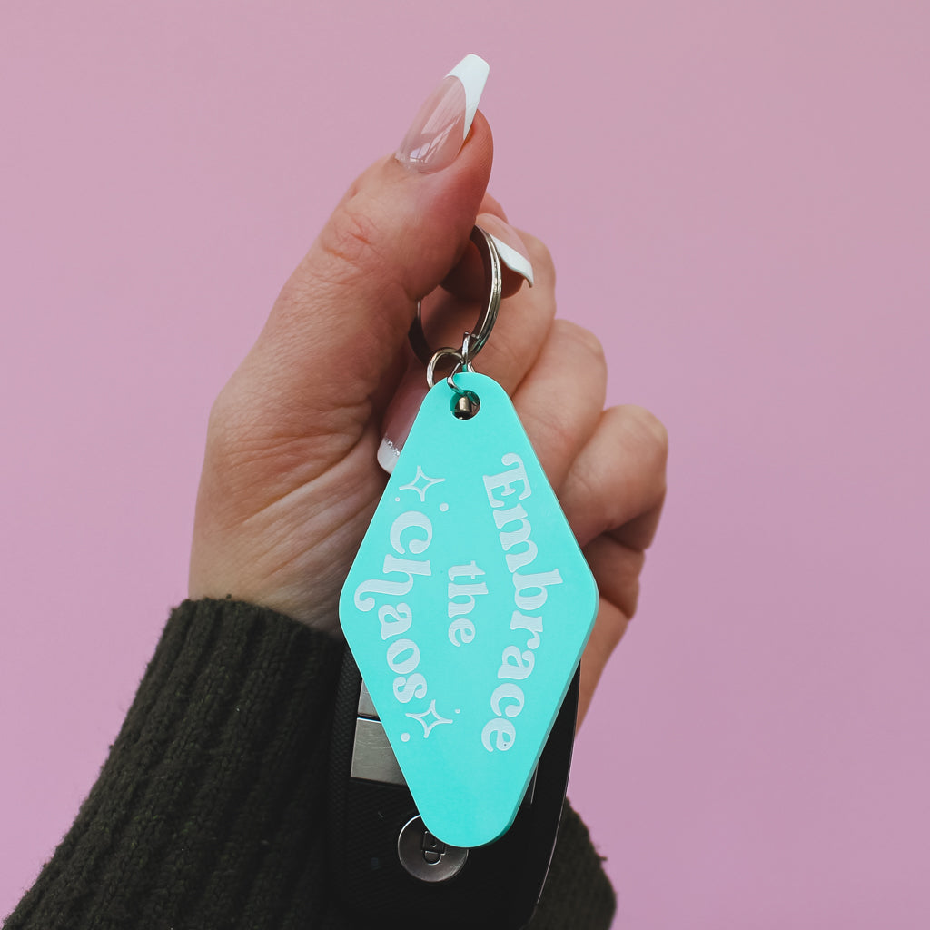 Hand holding a turquoise motel style keyring with 'Embrace the Chaos' text against a pink background