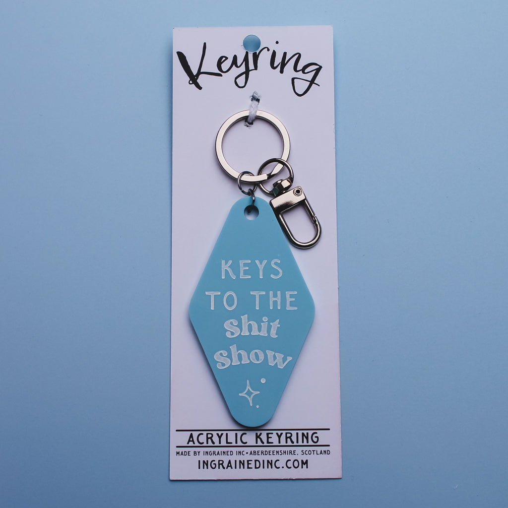 Blue motel keychain saying 'keys to the shit show' on a white backing  card against a blue background