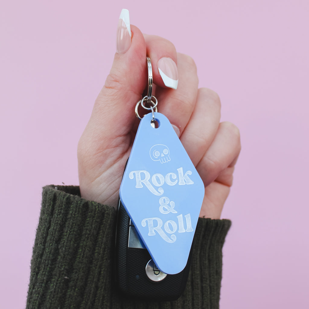 Hand holding a Motel style keychain with 'Rock & Roll' text against a pink background
