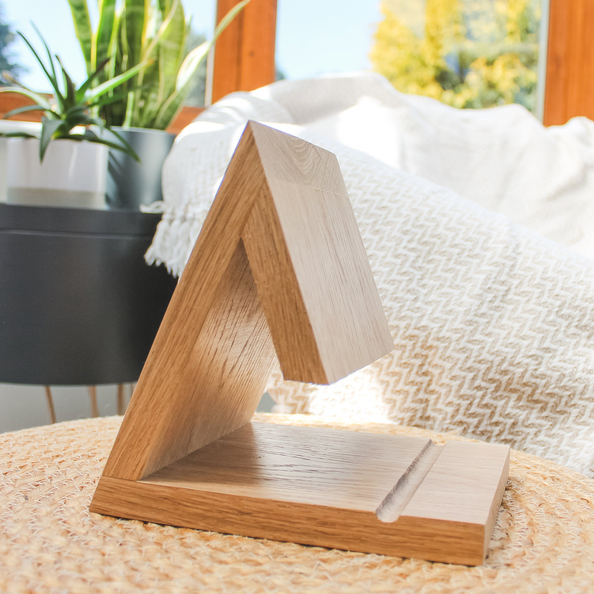 A triangular wooden display stand made of light oak wood, shown in a cozy, sunlit home setting on a woven rug with a patterned throw blanket and a potted plant in the background.