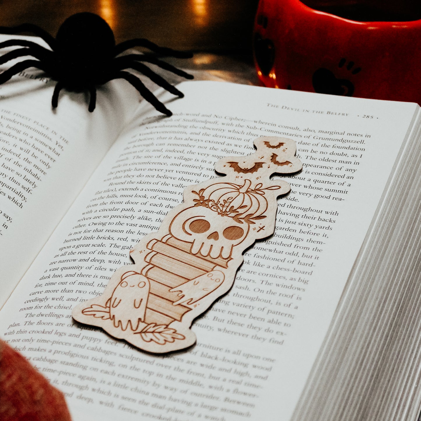 Wooden engraved bookmark with halloween design, including a pumpkin, skull and ghosts, placed on a book with spooky decorations placed around 