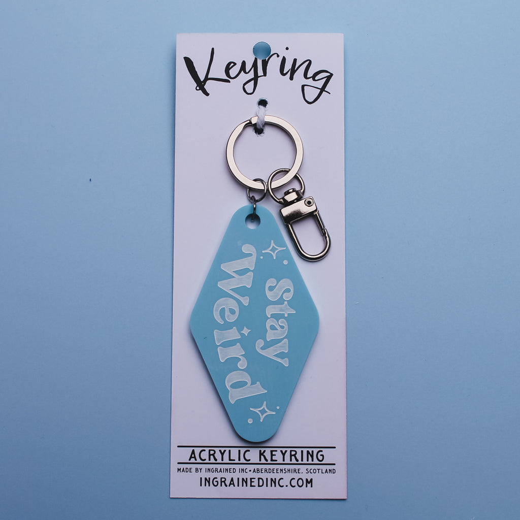 Blue motel style acrylic keyring with 'Stay Weird' text on a white  backing 
card against a blue background