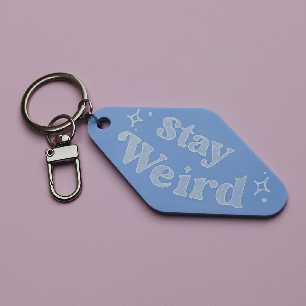 Blue motel style keyring tag with 'Stay Weird' text on a pink background