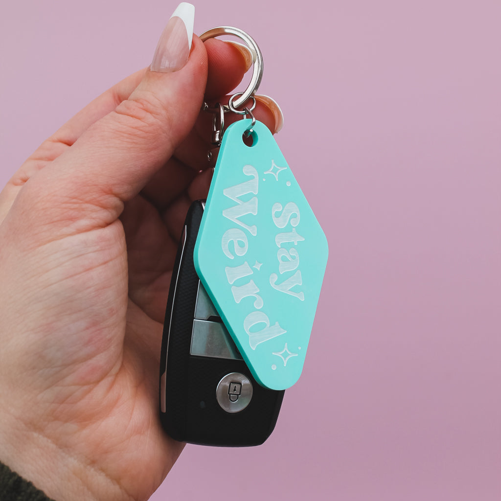 Hand holding a motel style keychain with a turquoise tag that says 'Stay Weird' against a pink background