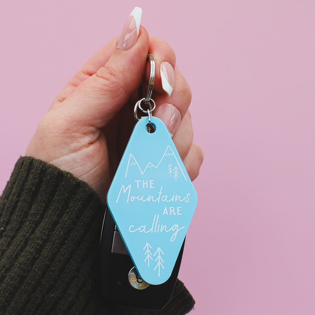 Hand holding a blue motel style keychain with 'The Mountains Are Calling' text against a pink background