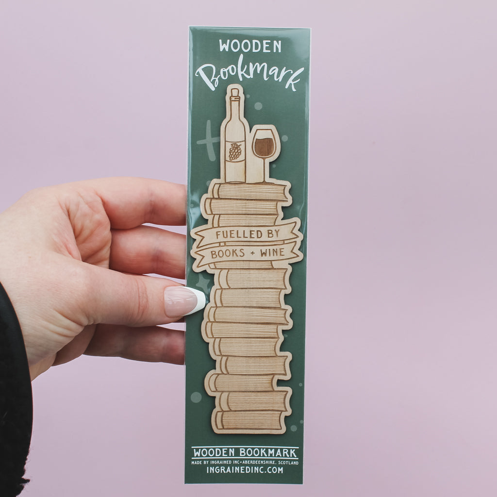 Wooden bookmark with books and wine bottle design in its packaging held by a hand against a light pink background
