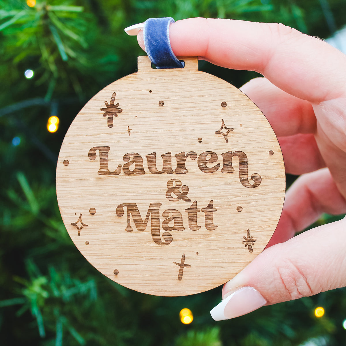 Wooden ornament with a couples name  engraved, held against a Christmas tree background.