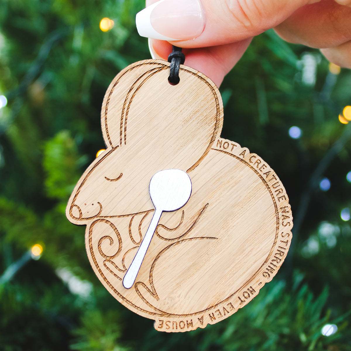 Wooden sleeping mouse xmas ornament with engraved design and text, held against a Christmas tree background.