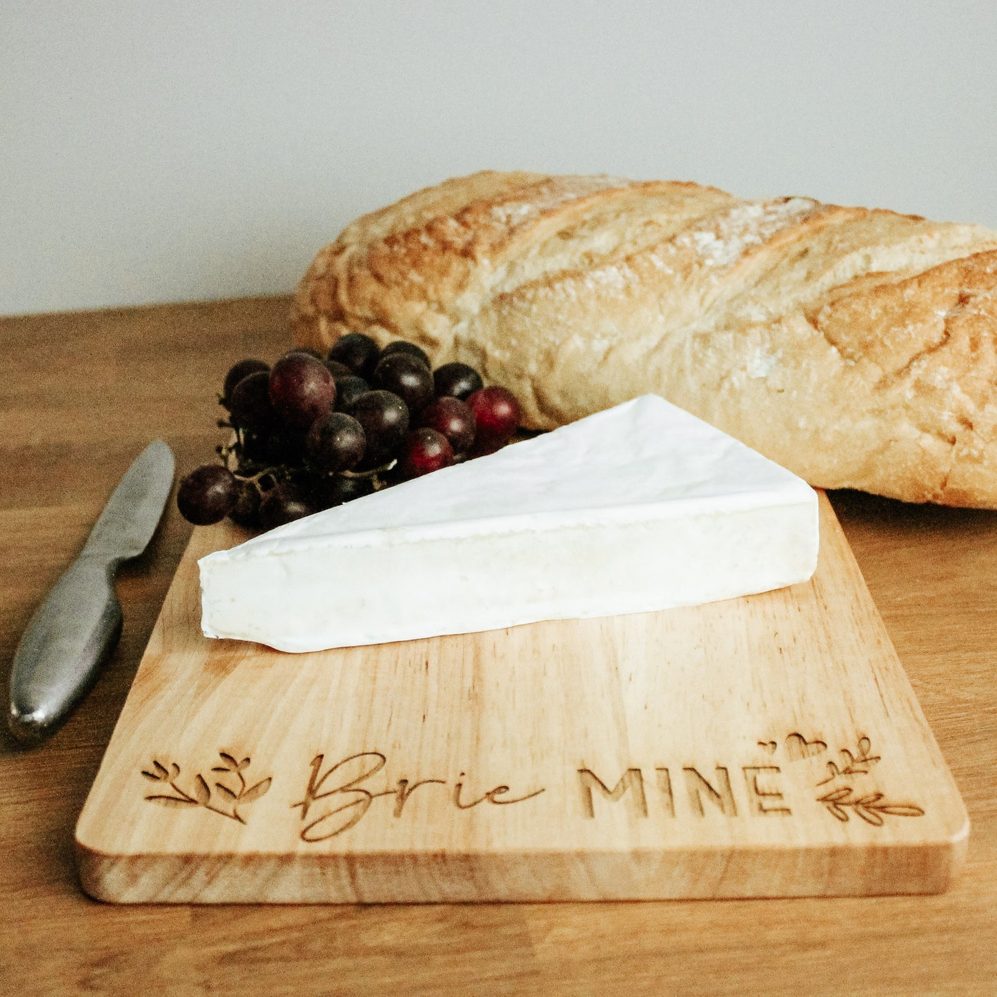 Rustic light wood chopping board with the words brie mine engraved on it with a botanical design on either side of the text. The board has a triangle of brie on it with some grapes and a loaf of bread at teh side