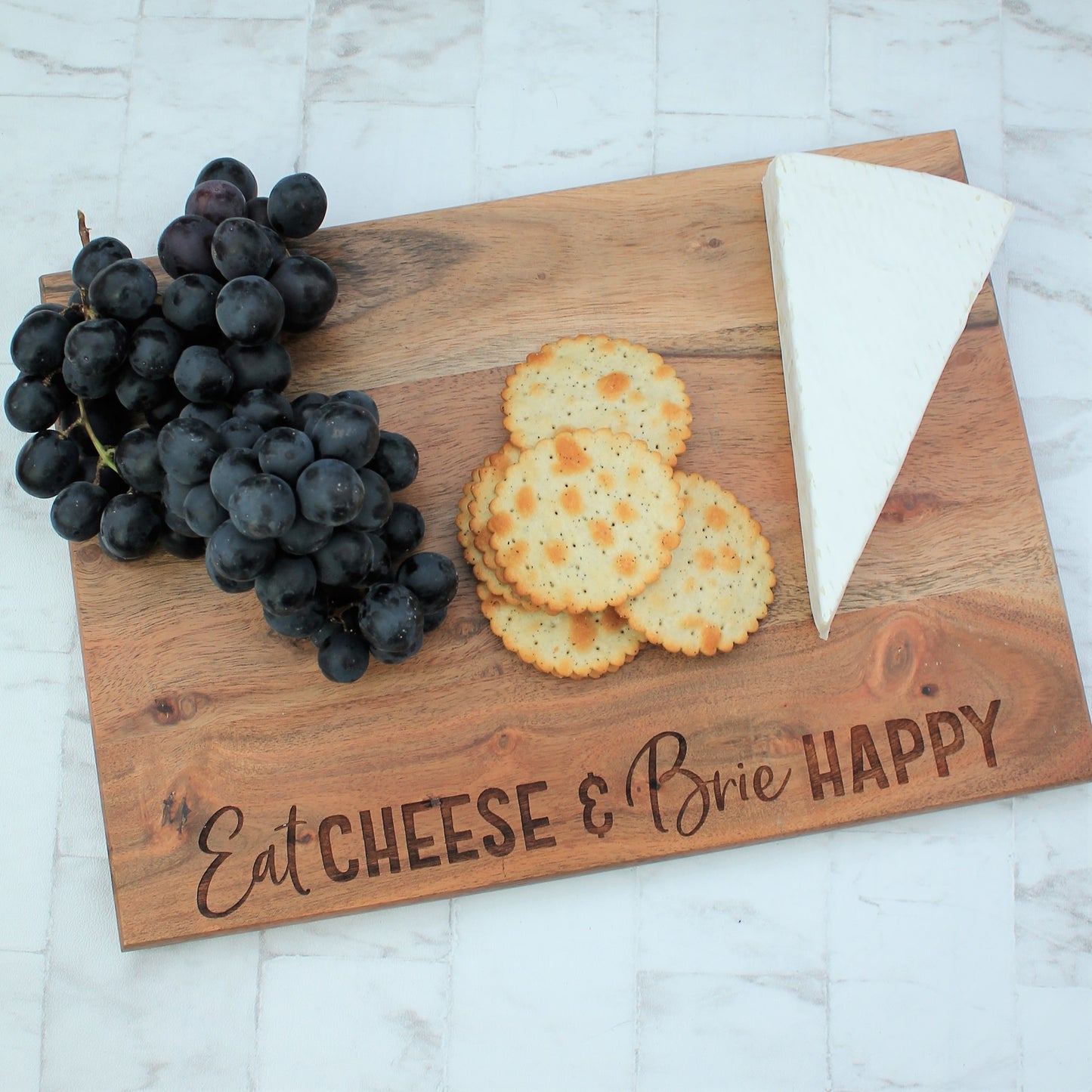 Wooden engraved chopping board with the words eat cheese and brie happy. With brie, grapes and crackers on top