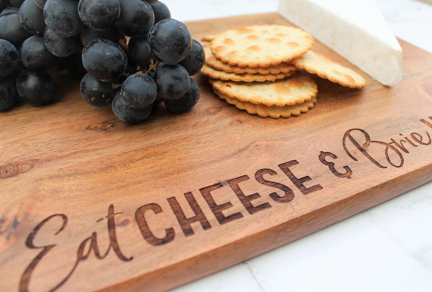 wooden cheese board with brie cracker and graes on top. engraved with the words eat cheese and brie happy