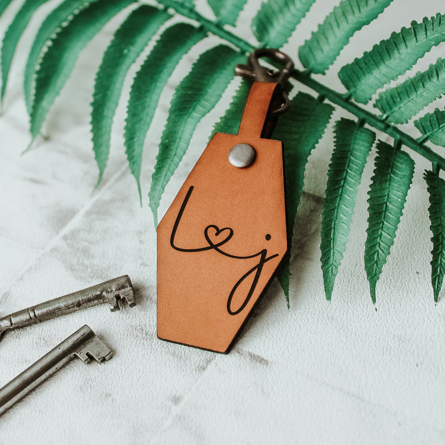 whisky coloured brown leather keyring with initials engraved on it connected by a heart design. with a swivel clasp attached to clip onto keys or a bag